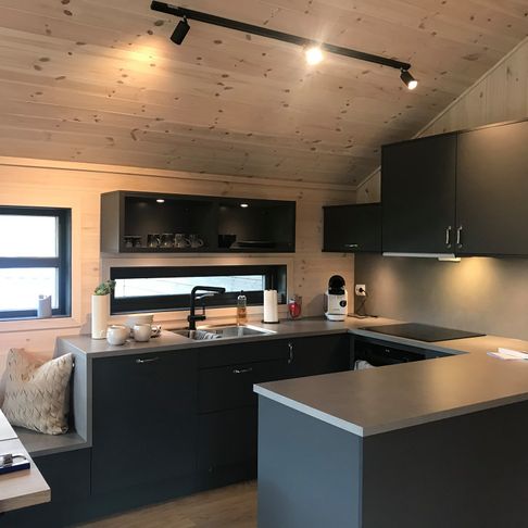 Et grått lite kjøkken med mye god naturlig lys fra vinduene samt lysene som er installert under kjøkkenskapene og i taket